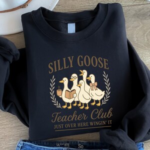 May include: Black sweatshirt featuring a cartoon goose design with the text "Silly Goose Teacher Club" and "Just Over Here Wingin' It". The graphic includes geese with a book, bow, and coffee cup.