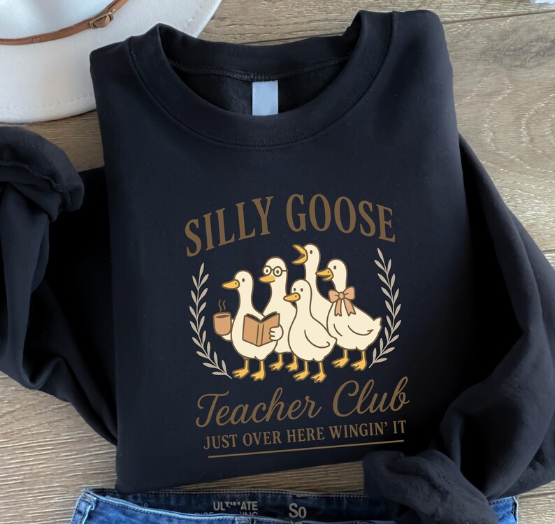 May include: Black sweatshirt featuring a cartoon goose design with the text "Silly Goose Teacher Club" and "Just Over Here Wingin' It". The graphic includes geese with a book, bow, and coffee cup.