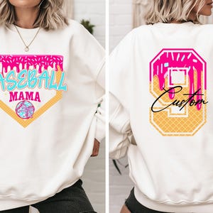 May include: White sweatshirt with a pink, blue, and yellow baseball-themed graphic on the front that says "Baseball Mama". The back of the sweatshirt has a pink, yellow, and white number 9 with the word "Custom" written in black.