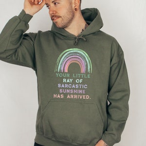 Puede incluir: Una sudadera con capucha verde con un gr&aacute;fico de arco&iacute;ris y el texto "Your little ray of sarcastic sunshine has arrived."