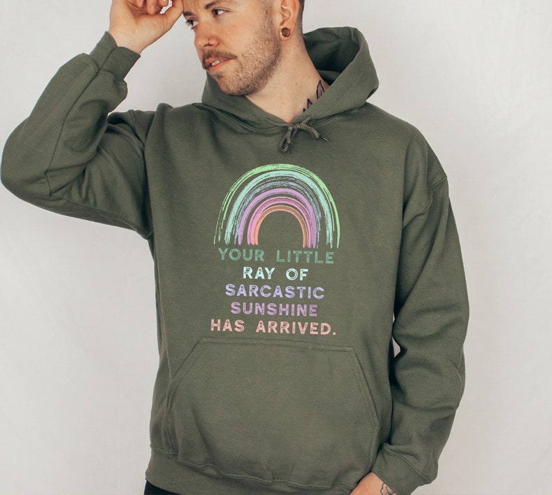 Puede incluir: Una sudadera con capucha verde con un gr&aacute;fico de arco&iacute;ris y el texto "Your little ray of sarcastic sunshine has arrived."