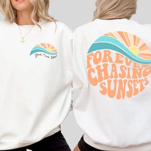 May include: White sweatshirt with a retro sunset graphic on the front and back. The front design includes a sun and waves with the text "Your Text Here". The back features the words "Forever Chasing Sunsets" in orange.