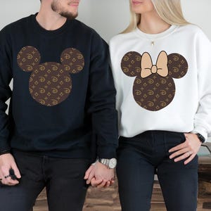 Puede incluir: Dos sudaderas, una azul marino y otra blanca, cada una con un dise&ntilde;o de cabeza de Mickey Mouse. Las cabezas de Mickey Mouse est&aacute;n rellenas con un patr&oacute;n marr&oacute;n y dorado. La sudadera blanca tiene un lazo beige.
