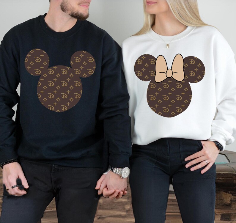 Puede incluir: Dos sudaderas, una azul marino y otra blanca, cada una con un dise&ntilde;o de cabeza de Mickey Mouse. Las cabezas de Mickey Mouse est&aacute;n rellenas con un patr&oacute;n marr&oacute;n y dorado. La sudadera blanca tiene un lazo beige.