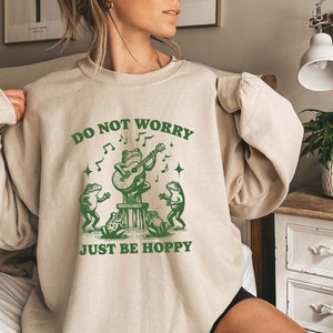 May include: A beige sweatshirt with a green graphic of frogs playing music. The text on the sweatshirt reads "Do Not Worry Just Be Hoppy".