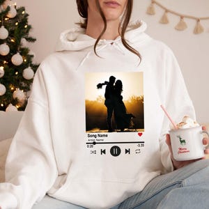 May include: White hoodie featuring a graphic of a couple embracing at sunset, designed to resemble a music player interface. The hoodie also has a mug with a deer and the text "Merry Christmas".
