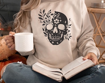 May include: A beige sweatshirt featuring a black floral skull design. The skull is decorated with leaves and flowers. The person in the image is holding a white mug and a book.