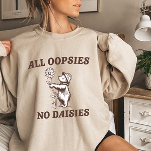 May include: A beige sweatshirt with a brown graphic of a bear holding a sunflower. The text "ALL OOPSIES NO DAISIES" is printed on the sweatshirt.