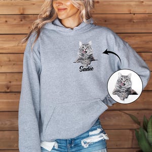 Puede incluir: Una sudadera con capucha gris claro con un retrato de gato personalizado y el nombre "Sadie" impreso debajo. El gato es gris y blanco con la lengua fuera. Tambi&eacute;n se muestra una imagen circular del gato.