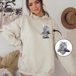Puede incluir: Sudadera con capucha color crema con un gr&aacute;fico de gato y el nombre "Sadie" impreso debajo. El gato es gris y blanco, con la lengua fuera. La sudadera tiene un bolsillo delantero y una capucha con cord&oacute;n.