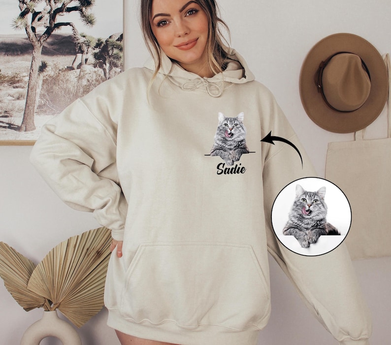 Puede incluir: Sudadera con capucha color crema con un gr&aacute;fico de gato y el nombre "Sadie" impreso debajo. El gato es gris y blanco, con la lengua fuera. La sudadera tiene un bolsillo delantero y una capucha con cord&oacute;n.