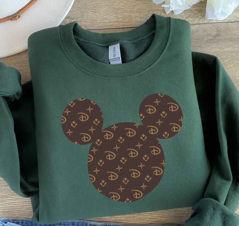 Puede incluir: Una sudadera verde oscuro con un dise&ntilde;o de Mickey Mouse. La silueta de Mickey Mouse est&aacute; rellena con un patr&oacute;n marr&oacute;n de logotipos. La sudadera est&aacute; doblada sobre una superficie de madera.