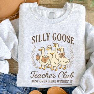 May include: Pale grey sweatshirt featuring the words "SILLY GOOSE Teacher Club" and a graphic of five cartoon ducks. One duck wears glasses and holds a book, another holds a coffee cup. The phrase "JUST OVER HERE WINGIN' IT" is below.