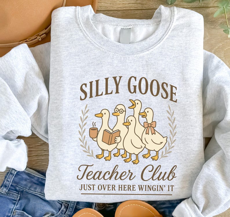 May include: Pale grey sweatshirt featuring the words "SILLY GOOSE Teacher Club" and a graphic of five cartoon ducks. One duck wears glasses and holds a book, another holds a coffee cup. The phrase "JUST OVER HERE WINGIN' IT" is below.