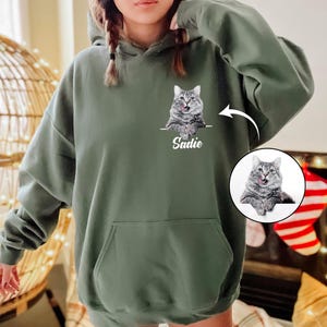 Op de afbeelding: Een olijfgroene hoodie met een aangepast kattenportret en de naam "Sadie" eronder. De kat is grijs en wit met zijn tong uit. De hoodie heeft een voorzak en een capuchon met trekkoord.