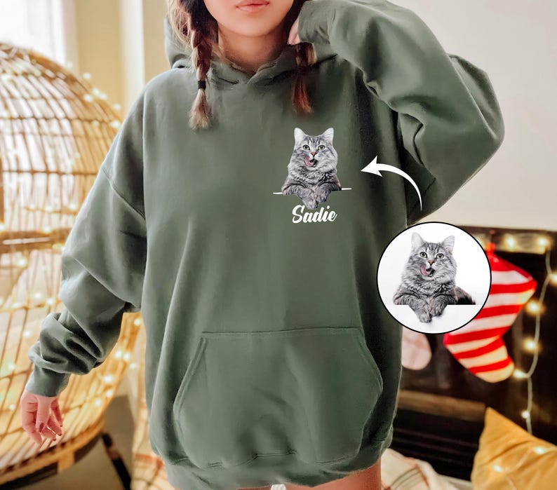 Op de afbeelding: Een olijfgroene hoodie met een aangepast kattenportret en de naam "Sadie" eronder. De kat is grijs en wit met zijn tong uit. De hoodie heeft een voorzak en een capuchon met trekkoord.