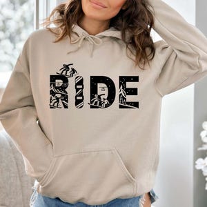 May include: A beige hoodie with a front pocket and a drawstring hood. The word "RIDE" is printed in black, with each letter featuring a unique winter sports design, including snowboarders and a snowy mountain scene.