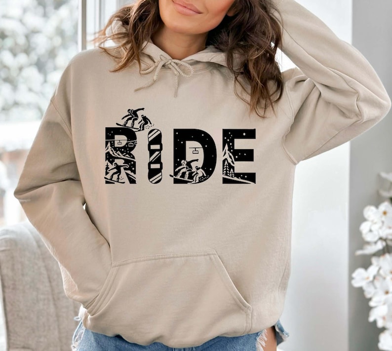 May include: A beige hoodie with a front pocket and a drawstring hood. The word "RIDE" is printed in black, with each letter featuring a unique winter sports design, including snowboarders and a snowy mountain scene.