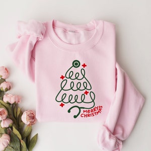 Pu&ograve; includere: Felpa rosa chiaro con un disegno di albero di Natale realizzato con uno stetoscopio verde scuro e croci rosse. Le parole "MERRY CHRISTMAS" sono in rosso. Il capo &egrave; piegato su una superficie bianca.
