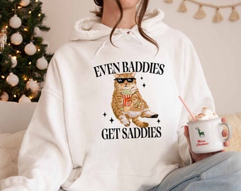 May include: White hoodie with a graphic of a cat wearing sunglasses and holding a bucket of popcorn. The text "Even Baddies Get Saddies" is printed on the hoodie.