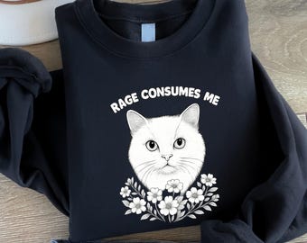 May include: Black sweatshirt featuring a white cat face with the text "RAGE CONSUMES ME" above it. The cat is surrounded by a floral wreath. The sweatshirt is folded, with a portion of blue jeans visible at the bottom.