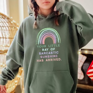 Puede incluir: Una sudadera con capucha verde con un gr&aacute;fico de arco&iacute;ris y el texto "Your little ray of sarcastic sunshine has arrived."