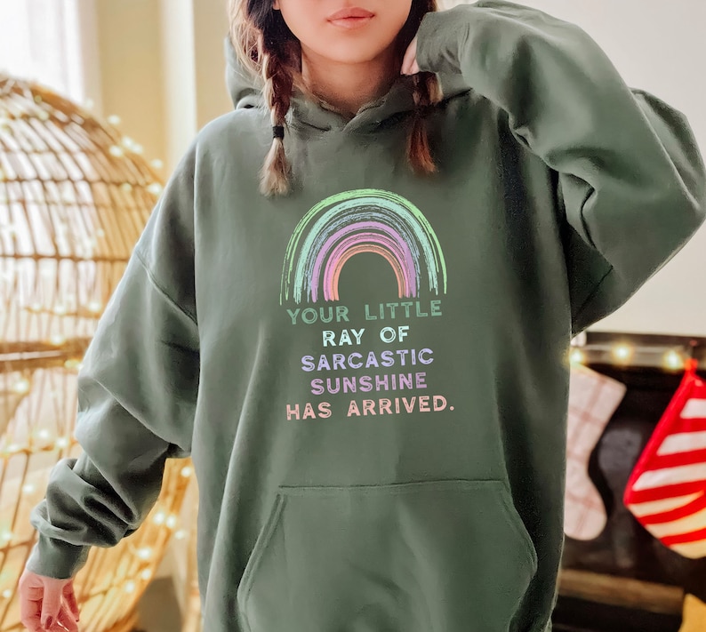 Puede incluir: Una sudadera con capucha verde con un gr&aacute;fico de arco&iacute;ris y el texto "Your little ray of sarcastic sunshine has arrived."