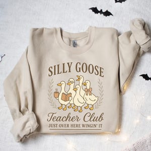 May include: A beige sweatshirt featuring the text "Silly Goose Teacher Club" and a graphic of five cartoon geese. One goose holds a coffee cup, another a book, and one wears glasses and a bow. The text "Just Over Here Wingin' It" is below.