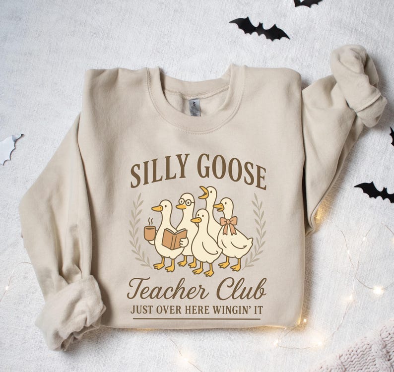 May include: A beige sweatshirt featuring the text "Silly Goose Teacher Club" and a graphic of five cartoon geese. One goose holds a coffee cup, another a book, and one wears glasses and a bow. The text "Just Over Here Wingin' It" is below.