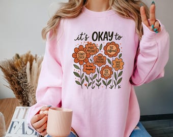 May include: A light pink sweatshirt with the text "it's OKAY to" above a floral design. The flowers have phrases like "Make Mistakes" and "Have Bad Days". The sweatshirt is a comfortable, casual style.