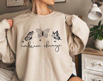 May include: A beige sweatshirt with a graphic of three butterflies and the text "embrace change" printed on the front.