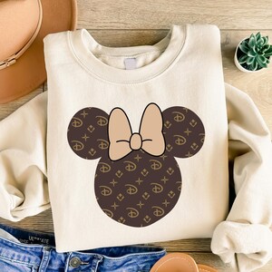 Puede incluir: Sudadera de color crema con un dise&ntilde;o de Minnie Mouse. El dise&ntilde;o incluye un lazo marr&oacute;n y orejas de rat&oacute;n marrones con un logotipo repetido. La sudadera est&aacute; hecha de un material suave y tiene mangas largas.
