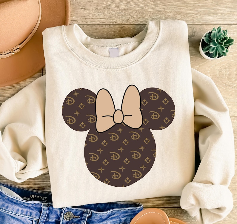 Puede incluir: Sudadera de color crema con un dise&ntilde;o de Minnie Mouse. El dise&ntilde;o incluye un lazo marr&oacute;n y orejas de rat&oacute;n marrones con un logotipo repetido. La sudadera est&aacute; hecha de un material suave y tiene mangas largas.