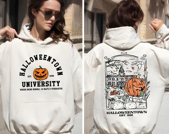 May include: Cream-colored Halloween-themed hoodie with a front graphic of a jack-o'-lantern and the text "Halloweentown University." The back features a detailed illustration of a spooky town scene with a pumpkin. The hoodie has a hood and a front pocket.