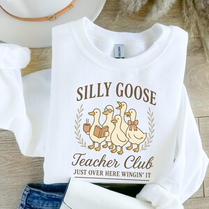 May include: White sweatshirt featuring the text "SILLY GOOSE Teacher Club" and a graphic of five cartoon geese. One goose holds a coffee cup, another a book, and one wears glasses and a bow. The phrase "JUST OVER HERE WINGIN' IT" is below the graphic.