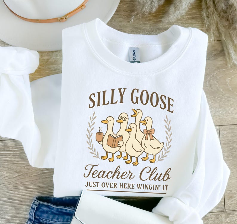 May include: White sweatshirt featuring the text "SILLY GOOSE Teacher Club" and a graphic of five cartoon geese. One goose holds a coffee cup, another a book, and one wears glasses and a bow. The phrase "JUST OVER HERE WINGIN' IT" is below the graphic.