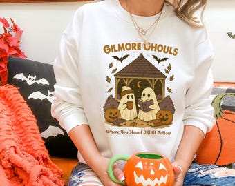 May include: White sweatshirt with a Halloween-themed graphic. The design features two ghost characters in a gazebo, pumpkins, bats, and the text "GILMORE GHOULS Where You Haunt I Will Follow". The sweatshirt is a cozy, seasonal apparel item.