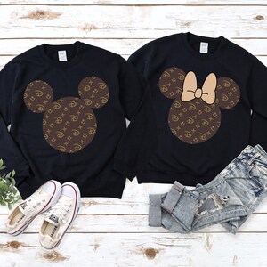 Puede incluir: Dos sudaderas negras con dise&ntilde;os de Mickey Mouse. Una presenta la silueta cl&aacute;sica de Mickey, la otra un lazo. Ambos dise&ntilde;os est&aacute;n rellenos con un patr&oacute;n marr&oacute;n y dorado. Zapatillas blancas y pantalones cortos de mezclilla tambi&eacute;n est&aacute;n en la imagen.