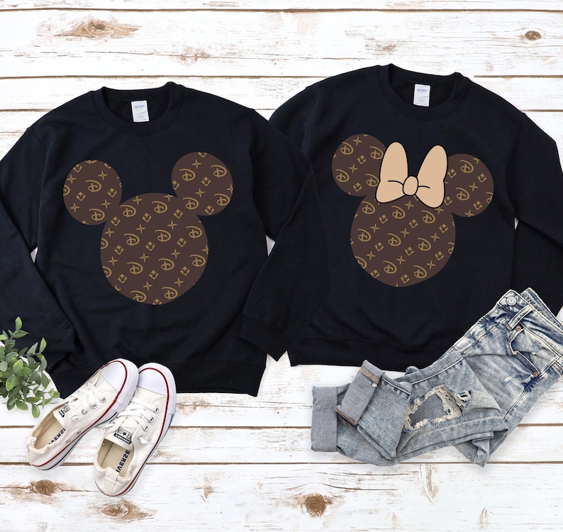 Puede incluir: Dos sudaderas negras con dise&ntilde;os de Mickey Mouse. Una presenta la silueta cl&aacute;sica de Mickey, la otra un lazo. Ambos dise&ntilde;os est&aacute;n rellenos con un patr&oacute;n marr&oacute;n y dorado. Zapatillas blancas y pantalones cortos de mezclilla tambi&eacute;n est&aacute;n en la imagen.