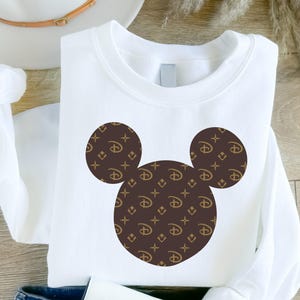 Puede incluir: Sudadera blanca con un dise&ntilde;o de Mickey Mouse. La silueta de Mickey Mouse est&aacute; compuesta por tres c&iacute;rculos marrones, cada uno lleno con un patr&oacute;n repetido del logotipo de Disney y un logotipo 'LV' estilizado.