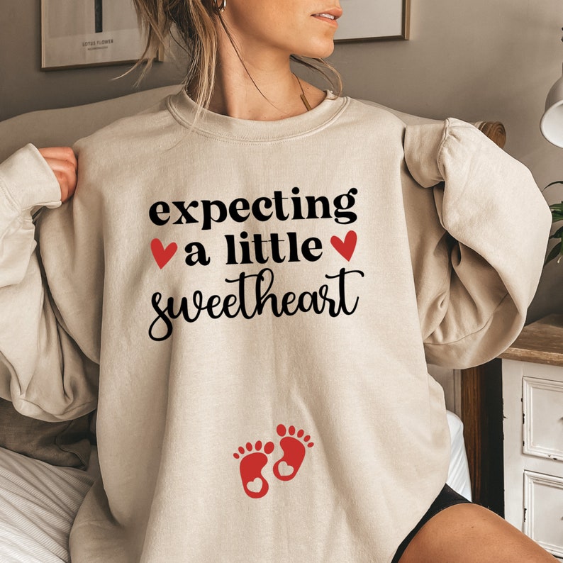 May include: A beige sweatshirt with the text "expecting a little sweetheart" and two red hearts. Below the text is a pair of red footprints with a red heart in each.