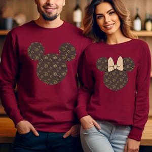 Puede incluir: Sudaderas burdeos de cuello redondo. Una presenta un dise&ntilde;o de cabeza de Mickey Mouse con un patr&oacute;n marr&oacute;n y dorado. La otra tiene un dise&ntilde;o de cabeza de Minnie Mouse con un lazo y el mismo patr&oacute;n. Las sudaderas las llevan dos personas.