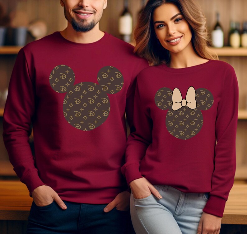Puede incluir: Sudaderas burdeos de cuello redondo. Una presenta un dise&ntilde;o de cabeza de Mickey Mouse con un patr&oacute;n marr&oacute;n y dorado. La otra tiene un dise&ntilde;o de cabeza de Minnie Mouse con un lazo y el mismo patr&oacute;n. Las sudaderas las llevan dos personas.