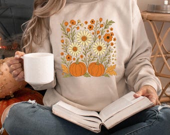 May include: Beige sweatshirt featuring a floral and pumpkin design. The graphic includes orange pumpkins, daisies, and other flowers in shades of orange, yellow, and green. The sweatshirt is a neutral color, suitable for fall.