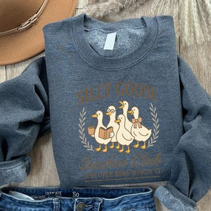 May include: A dark grey sweatshirt featuring the text "SILLY GOOSE Teachers Club" and a graphic of five cartoon ducks. One duck wears glasses and another a bow. The sweatshirt is made of a soft material.