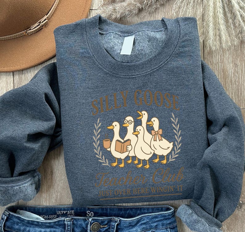 May include: A dark grey sweatshirt featuring the text "SILLY GOOSE Teachers Club" and a graphic of five cartoon ducks. One duck wears glasses and another a bow. The sweatshirt is made of a soft material.