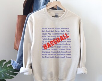 May include: Cream-colored sweatshirt with a list of baseball terms in blue and the word "BASEBALL" in red. The sweatshirt is hanging on a wooden hanger.