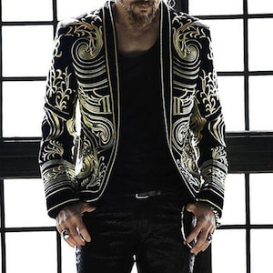 Puede incluir: Una chaqueta negra con bordados dorados con un patrón floral. La chaqueta tiene un cuello chal y un cierre de un solo botón.