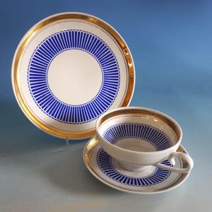 Peut inclure: Un service à thé blanc avec une tasse, une soucoupe et une assiette, orné d'un motif à rayures bleues et de bordures dorées. La tasse a une anse incurvée. L'ensemble est posé sur une surface bleu clair.