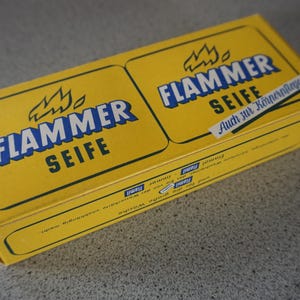 Könnte beinhalten: Eine gelbe Schachtel mit dem Text "FLAMMER SEIFE" und einer Flammengrafik. Die Schachtel hat auch den Text "Auch zur Kopfwasche" in Schwarz.